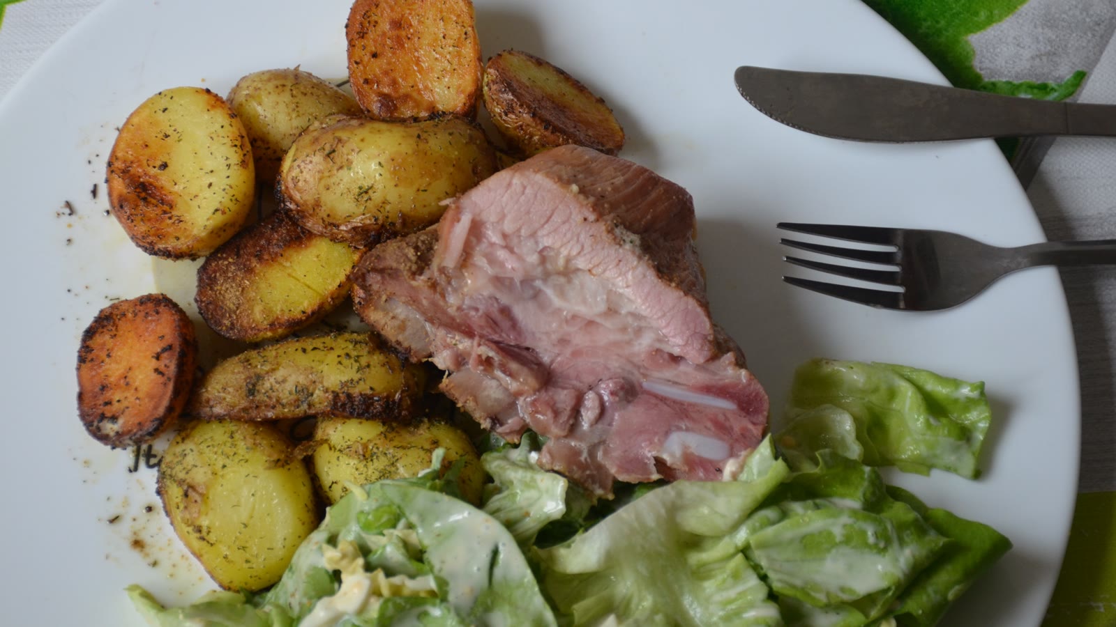 Skiveskåret hamburgerryg med lyserød midte, stegte kartofler og grøn salat på en hvid tallerken
